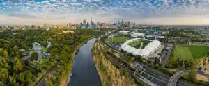 Aerial panoramic view of Melbourne CBD skyline with the Yarra River, Royal Botanic Gardens, AAMI Park, and Melbourne Park sports precinct under a cloudy sky